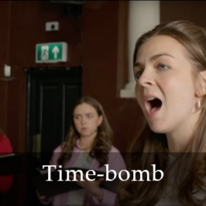 A photo of a white woman with brunette hair singing. Behind her is another white woman with brunette hair and between them a white pillar against dark red walls. "Time-bomb" is written in white at the bottom of the image.